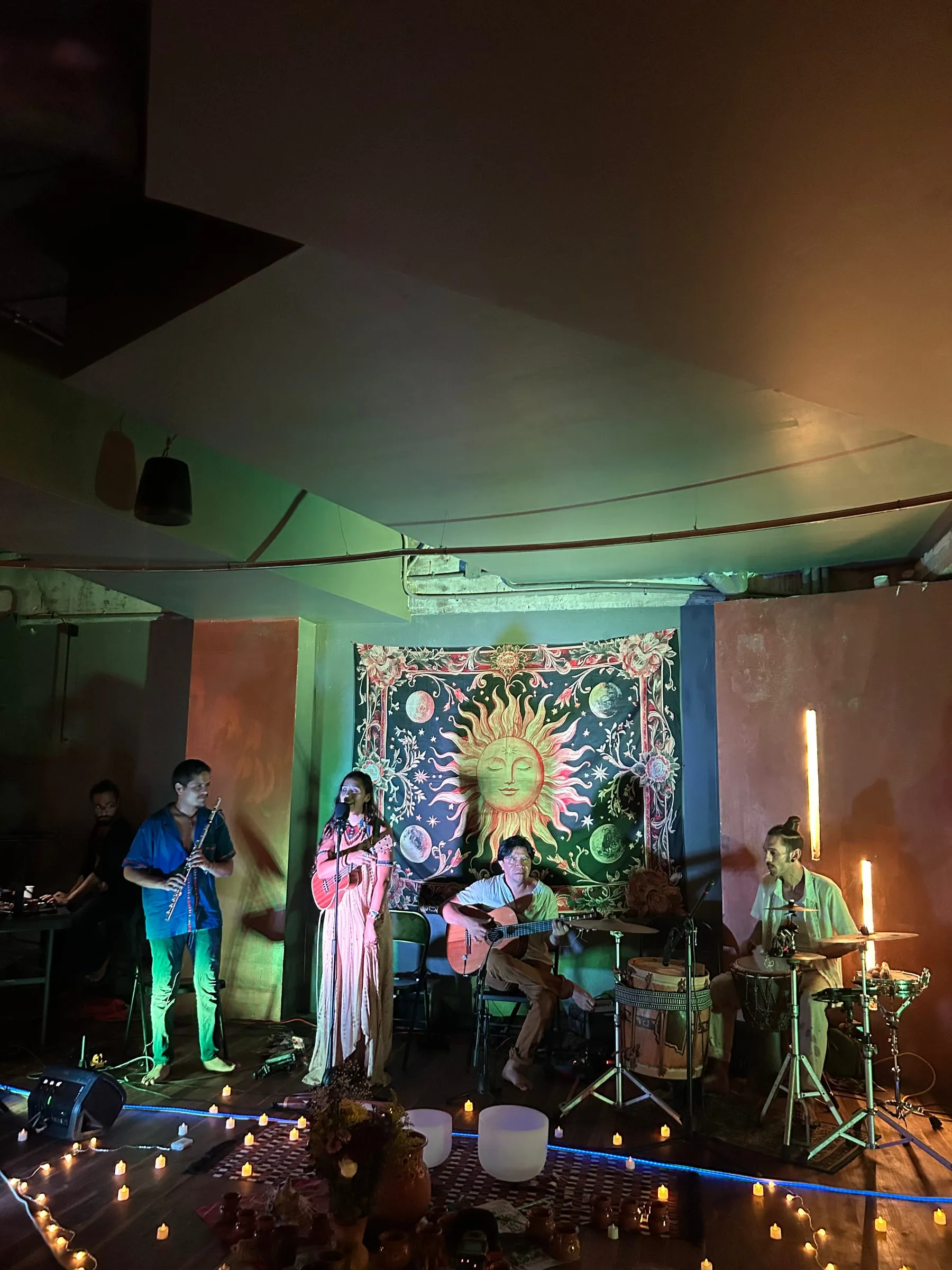 Sesión de música en vivo en Centro Calea: flauta, voz, guitarra y percusión frente a un tapiz del sol, iluminados por velas.